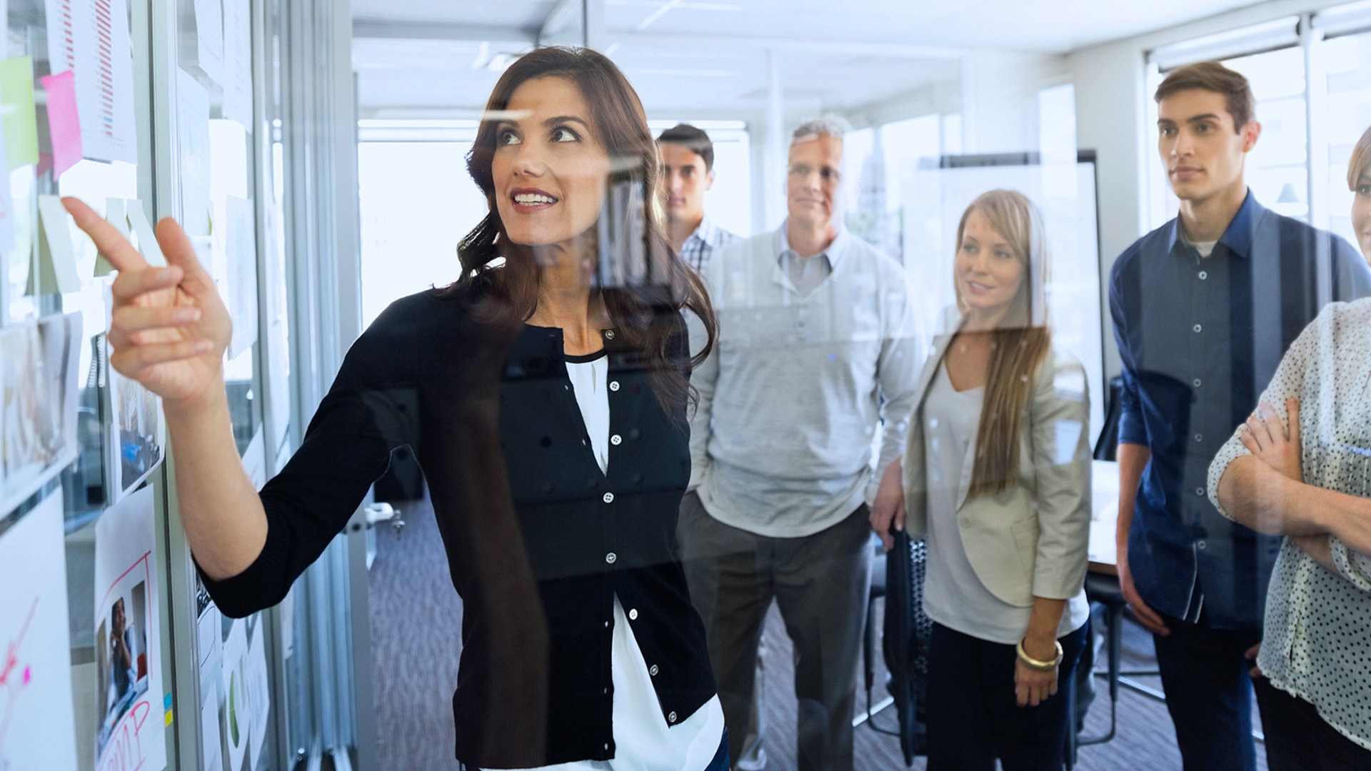 Eine Frau zeigt auf eine Tafel in einem hellen Büro und hält eine Präsentation vor fünf Kollegen, die aufmerksam um sie herum versammelt sind. Bei den Kollegen handelt es sich um eine Mischung aus Männern und Frauen in Berufskleidung, die vor einem großen Fenster stehen.