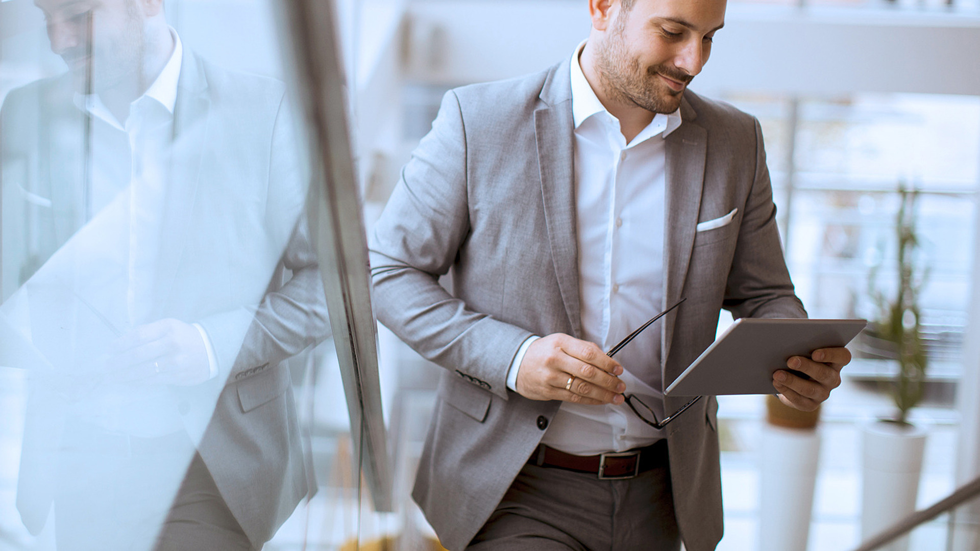 Ein Mann im grauen Anzug hält ein Tablet und eine Brille in der Hand und geht eine helle, moderne Treppe hinauf. Ein großes Fenster spiegelt sein Bild. Er ist auf das Tablet konzentriert und lächelt leicht, was darauf hindeutet, dass er sich mit dem Inhalt beschäftigt.